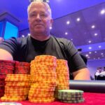 Ein Tisch mit einem Kartenspiel, Pokerchips und Getränken, umgeben von vier Personen, die konzentriert Poker spielen.