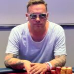 Ein Mann und eine Frau sitzen an einem Pokertisch und spielen gegeneinander. Der Mann hat eine Hand mit Karten in der Luft, während die Frau konzentriert ihre Karten betrachtet. Im Hintergrund sind