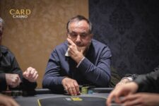Ein Mann mit dunklen Haaren und einem blauen Hemd sitzt an einem Pokertisch und hält Karten in der Hand. Er hat eine ernste Miene und schaut konzentriert auf seine Karten.