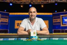 Ein Mann, der David Shmuel heißt, sitzt an einem Pokertisch und betrachtet konzentriert seine Karten. Er hat kurze dunkle Haare und trägt ein weißes Hemd. Im Hinter