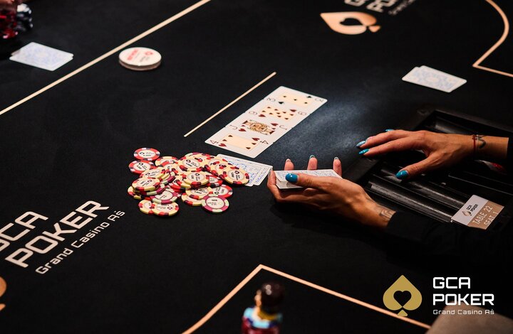 Ein Mann sitzt an einem Pokertisch mit Chips und Karten vor sich. Er trägt eine schwarze Kappe und konzentriert sich auf sein Blatt. Im Hintergrund sind weitere Spieler und ein Dealer zu sehen.
