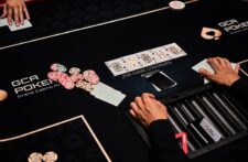 Ein Poker-Tisch mit Spielkarten, Chips und Getränken, umgeben von mehreren Personen, die konzentriert spielen.