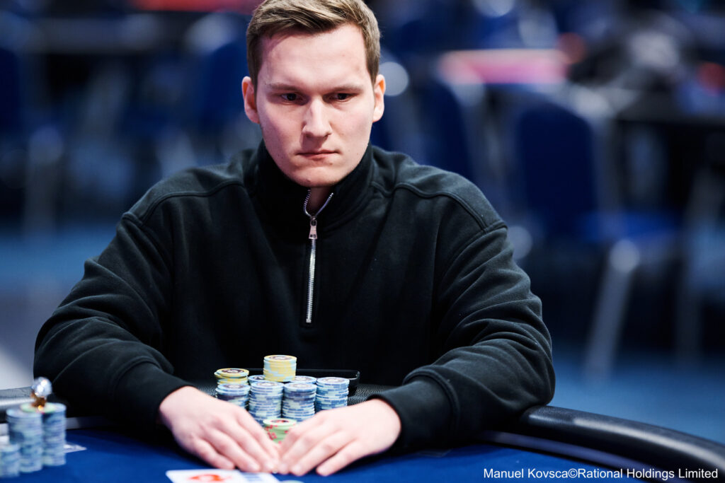 Ein Mann sitzt an einem Pokertisch und betrachtet konzentriert seine Karten. Im Hintergrund sind andere Spieler und ein Dealer zu sehen.