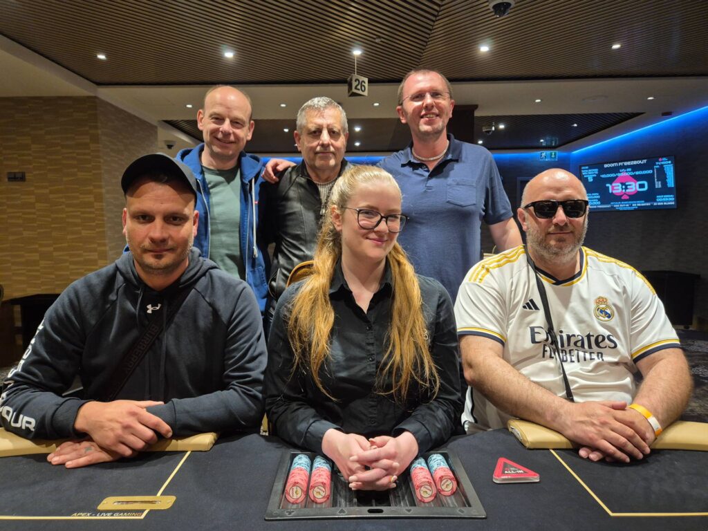 Ein Gruppenbild von fünf Personen, die an einem Pokertisch sitzen und Karten halten. Die Spieler haben konzentrierte Gesichtsausdrücke, während sie an einem Freezeout-Turnier teilnehmen.