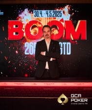 Ein Mann mit dunklem Haar und Bart sitzt an einem Pokertisch, konzentriert auf seine Karten. Er trägt ein dunkles Hemd und hat eine Pokerkarte in der Hand. Die Atmosphäre ist ged
