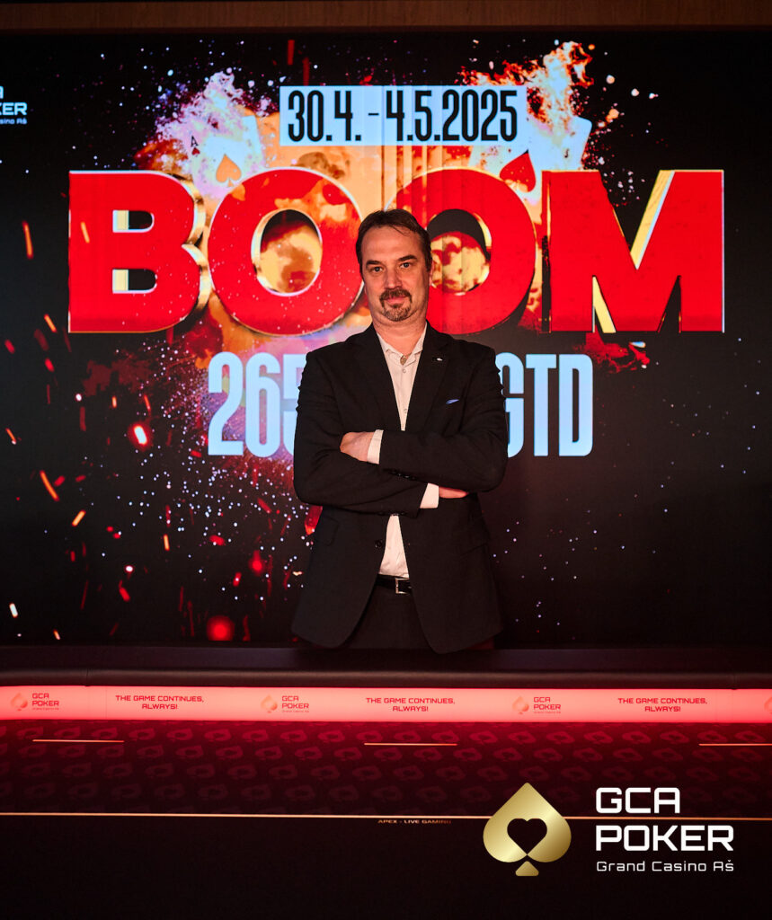 Ein Mann mit dunklem Haar und Bart sitzt an einem Pokertisch, konzentriert auf seine Karten. Er trägt ein dunkles Hemd und hat eine Pokerkarte in der Hand. Die Atmosphäre ist ged