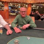 Ein Mann in einem Anzug und mit einer Sonnenbrille sitzt an einem Pokertisch und hält eine Hand mit Spielkarten. Ein Stapel Jetons liegt vor ihm.