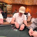 Ein Mann und eine Frau sitzen an einem Pokertisch und spielen eine Partie Poker. Der Mann hält Karten in der Hand, während die Frau konzentriert auf ihre eigenen Karten schaut. Im Hintergrund sind Poker