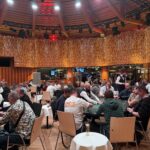 Ein Mann und eine Frau sitzen an einem Pokertisch, sie haben Karten in der Hand und schauen konzentriert auf den Tisch. Um sie herum sind Pokerchips und Getränke zu sehen.