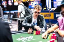 Ein Mann in einem dunklen Anzug und einer roten Krawatte sitzt an einem Pokertisch und betrachtet konzentriert seine Karten. Im Hintergrund sind andere Spieler zu sehen.