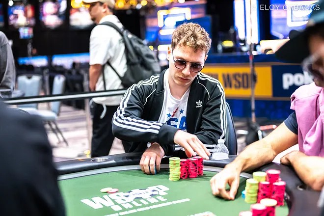 Ein Mann in einem dunklen Anzug und einer roten Krawatte sitzt an einem Pokertisch und betrachtet konzentriert seine Karten. Im Hintergrund sind andere Spieler zu sehen.