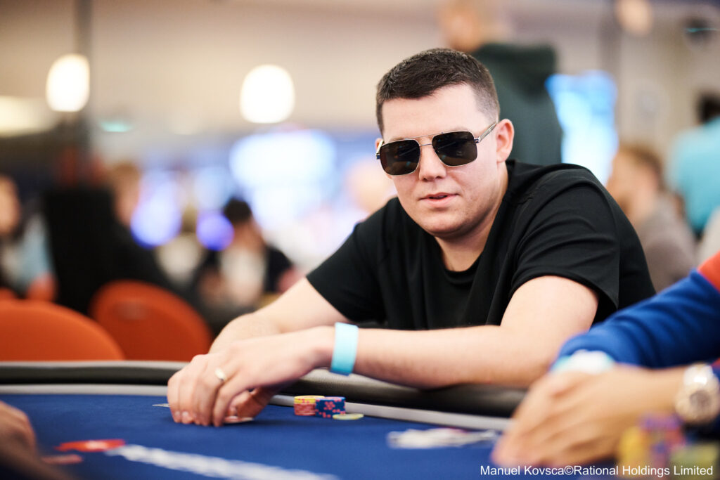 Ein Mann mit dunklen Haaren und Bart sitzt an einem Pokertisch und betrachtet konzentriert seine Karten. Er trägt ein schwarzes Hemd und hat eine ernste Miene.