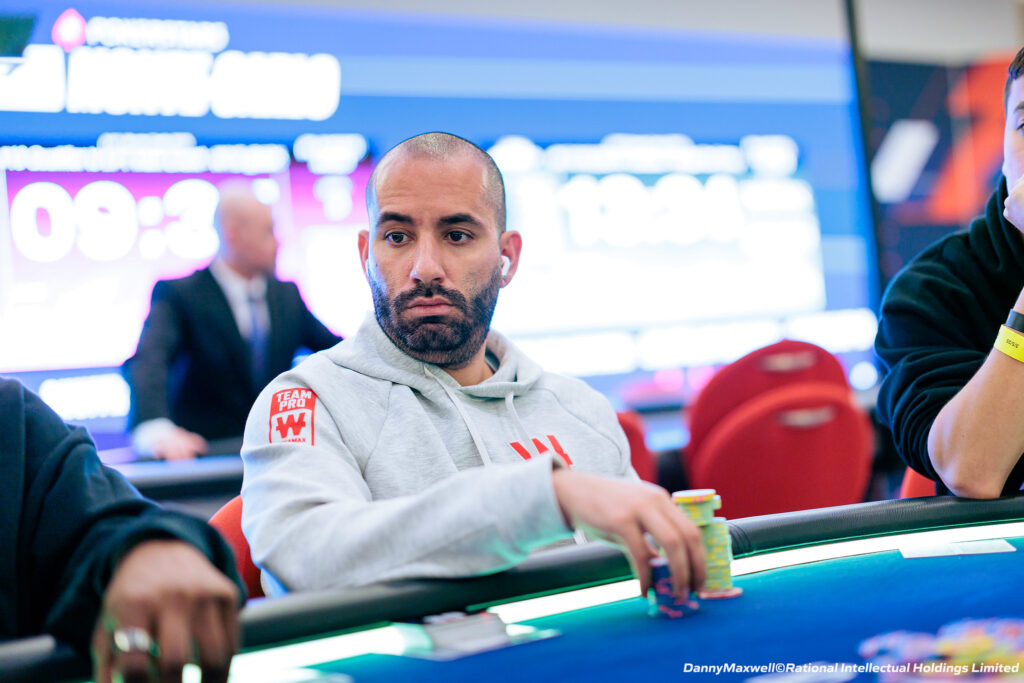Ein junger Mann mit dunklen Haaren und Bart, gekleidet in einem schwarzen Hemd und einer schwarzen Mütze, sitzt lächelnd an einem Pokertisch und hält Karten in der Hand.