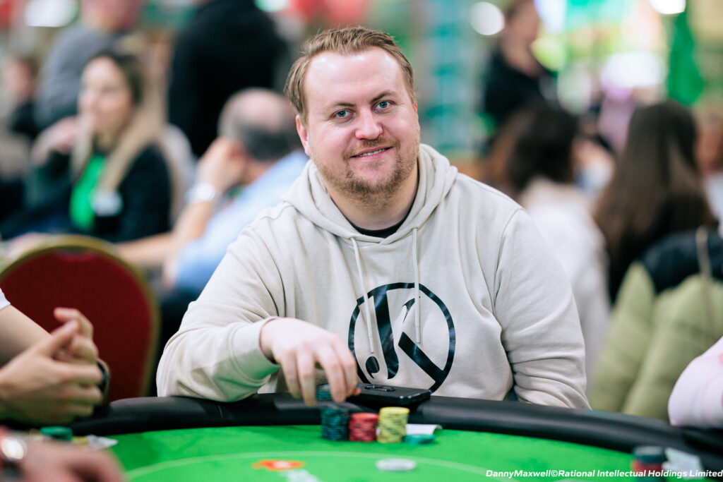 Ein Mann in schwarzem Anzug und roter Krawatte sitzt an einem Pokertisch und hält eine Hand mit Spielkarten. Im Hintergrund sind Chips und Gläser zu sehen.