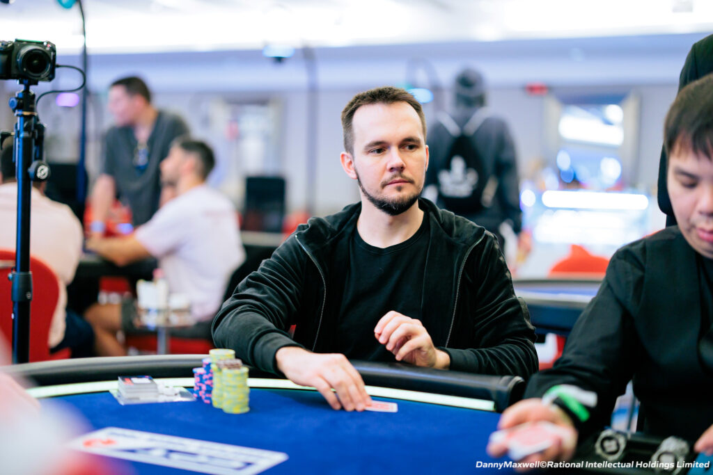 Ein Mann mit dunklen Haaren und Bart sitzt an einem Pokertisch und schaut konzentriert auf seine Karten. Er trägt ein schwarzes Hemd und eine schwarze Kappe. Im Hintergrund sind weitere Pok