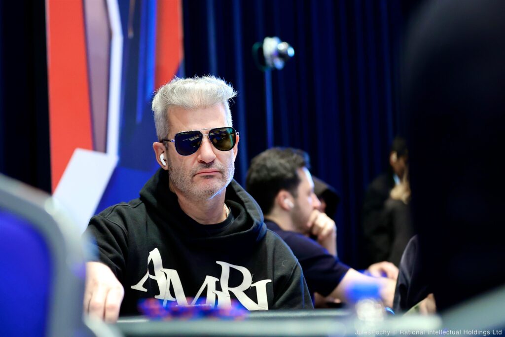 Ein Mann sitzt lächelnd an einem Pokertisch während eines Turniers. Er trägt ein schwarzes Shirt und hat eine Kappe auf dem Kopf. Im Hintergrund sind andere Spieler und Kartendealer zu sehen.