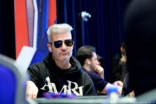 Ein Mann sitzt lächelnd an einem Pokertisch während eines Turniers. Er trägt ein schwarzes Shirt und hat eine Kappe auf dem Kopf. Im Hintergrund sind andere Spieler und Kartendealer zu sehen.