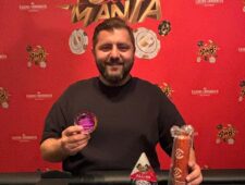 Ein Mann in schicker Kleidung und Sonnenbrille sitzt lässig an einem Pokertisch und hält Pokerchips in der Hand. Im Hintergrund sind andere Spieler und Spielkarten zu sehen.