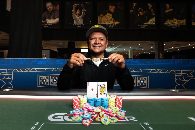Ein junger Mann sitzt an einem Pokertisch und hält zwei Spielkarten in der Hand. Er schaut konzentriert auf den Tisch, umgeben von Pokerchips und anderen Spielern im Hintergrund.