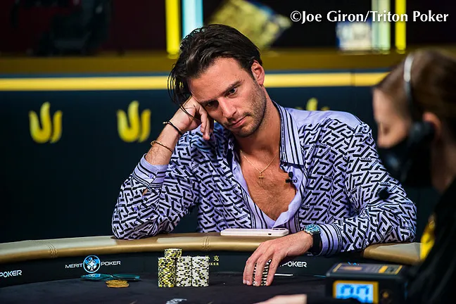 Ein Mann in einem dunkelblauen Anzug und einer Sonnenbrille sitzt an einem Pokertisch mit grünem Filz. Er betrachtet konzentriert seine Karten und seine Chips. Im Hintergrund sind
