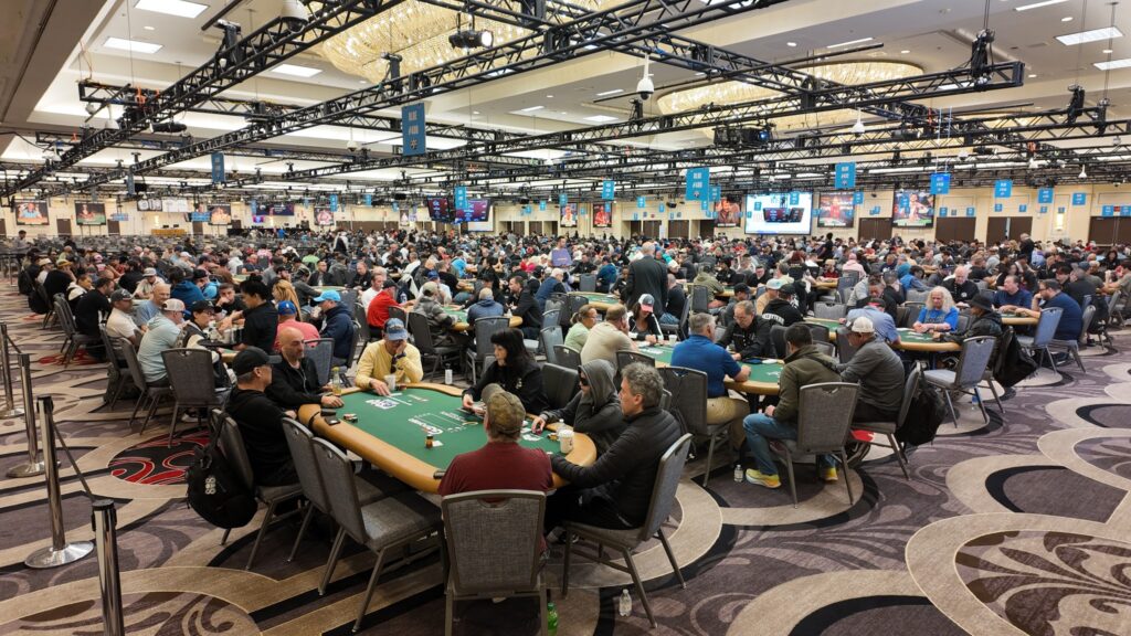 Ein professioneller Pokerspieler mit Sonnenbrille und Kopfhörern sitzt an einem Tisch während der World Series of Poker. Er hält Karten in der Hand und konzentriert sich auf das Spiel. Im H