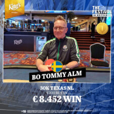 Ein Mann in einem schwarzen Anzug und einer schwarzen Sonnenbrille hält zwei Spielkarten in der Hand und schaut ernst in die Kamera. Im Hintergrund ist eine Casino-Umgebung mit Pokerchips und