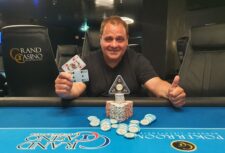 Ein Mann spielt Poker an einem Tisch in einem Casino. Er hält Karten in der Hand und setzt Pokerchips. Im Hintergrund sind weitere Spieler und Dealer zu sehen.