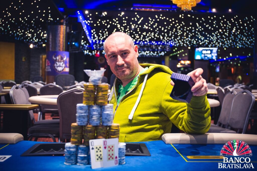 Ein Mann sitzt an einem Pokertisch in einem Casino, umgeben von anderen Spielern. Er hält mit ernstem Gesichtsausdruck Karten in der Hand und betrachtet sie konzentriert. Die Atmosph