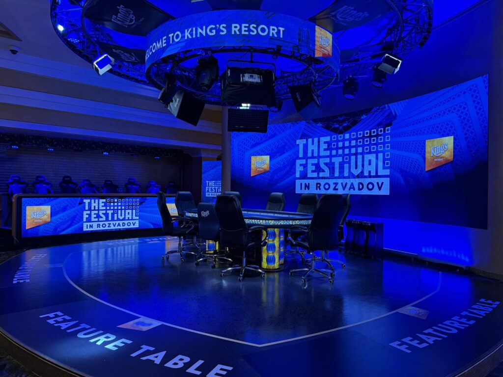 Ein Mann sitzt an einem Pokertisch mit Pokerchips und Karten vor sich. Er hat eine nachdenkliche Miene und scheint konzentriert auf sein Blatt zu schauen. Im Hintergrund sind weitere Spieler