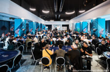 Ein Mann sitzt an einem Pokertisch und betrachtet seine Karten konzentriert, während er von anderen Spielern umgeben ist. Die Spannung und Intensität des Pokerspiels sind spürbar.