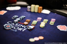 Ein Mann sitzt an einem Pokertisch und hält seine Karten in der Hand, während er konzentriert auf den Tisch schaut. Andere Spieler sind im Hintergrund zu sehen.