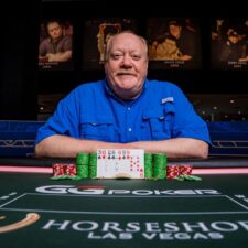 Ein älterer Mann mit Brille und grauem Haar sitzt an einem Pokertisch und hält eine Hand mit Spielkarten. Er trägt ein blaues Hemd und hat eine ernste Miene.
