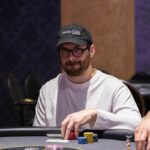 Ein Mann in einem schwarzen Anzug sitzt an einem Pokertisch und hält zwei Spielkarten in der Hand. Er schaut konzentriert auf die Karten und zeigt keine Emotionen. Im Hintergrund sind andere