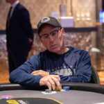 Ein Mann mit dunklen Haaren und einem blauen Hemd sitzt an einem Pokertisch und hält zwei Spielkarten in der Hand. Er schaut konzentriert auf den Tisch, umgeben von Chips und anderen Spiel