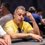 Ein Mann in einem schwarzen Anzug sitzt an einem Pokertisch und hält seine Karten mit ernstem Gesichtsausdruck. Um ihn herum sind Pokerchips und Getränke zu sehen.