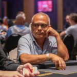 Ein glücklicher Mann mit Sonnenbrille und Kappe sitzt an einem Pokertisch und hält eine Hand mit Spielkarten. Andere Spieler und Chips sind im Hintergrund zu sehen.