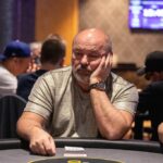 Ein Mann in einem schwarzen Anzug und einer Sonnenbrille sitzt am Pokertisch und hält Pokerchips in der Hand. Er wirkt konzentriert und ernsthaft.