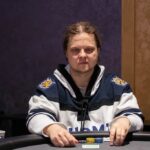 Ein Mann sitzt an einem Pokertisch und hält zwei Spielkarten in der Hand. Er trägt eine Kappe und konzentriert sich auf das Spiel. Im Hintergrund sind Pokerchips und andere Spieler zu sehen.