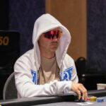 Ein Mann und eine Frau sitzen an einem Pokertisch und spielen Poker. Der Mann hat eine Hand mit Karten in der Hand, während die Frau konzentriert auf ihre Karten schaut. Im Hintergrund sind Pokerchips