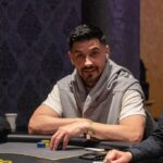 Ein Mann sitzt an einem Pokertisch und hält seine Spielkarten in der Hand. Er schaut konzentriert auf seine Karten, während die Chips vor ihm gestapelt sind. Im Hintergrund sind weitere Spieler und