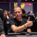 Ein Mann und eine Frau sitzen an einem Pokertisch und spielen eine Partie Poker. Der Mann hat eine Hand mit Karten in der Hand, während die Frau herausfordernd auf ihre Karten schaut. Im Hintergrund sind Poker