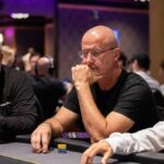 Ein Mann sitzt an einem Pokertisch und hält zwei Spielkarten in der Hand. Er schaut konzentriert auf den Tisch, umgeben von Pokerchips und anderen Spielern im Hintergrund.