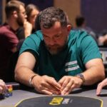 Ein Mann in einem Anzug und mit einer Sonnenbrille sitzt an einem Pokertisch und hält Karten in der Hand, während er konzentriert auf seine Gegner schaut.