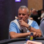 Ein Mann in einem schicken Anzug sitzt an einem Pokertisch, umgeben von Spielkarten, Jetons und einem Glas Whisky. Er betrachtet konzentriert seine Karten und wirkt entschlossen.