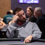 Ein Mann und eine Frau sitzen an einem Pokertisch und spielen Poker. Der Mann hat eine Hand aus Spielkarten, während die Frau ihre Karten betrachtet. Hinter ihnen sind Pokerchips und Getränke auf dem