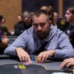 Ein glücklicher Mann mit einem breiten Lächeln sitzt an einem Pokertisch und hält eine Hand voll Spielkarten. Im Hintergrund sind andere Spieler und Pokerchips zu sehen.