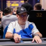 Ein Mann in einem eleganten Anzug sitzt an einem Pokertisch und hält eine Hand mit Spielkarten. Andere Spieler sind im Hintergrund zu sehen.