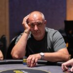 Ein Mann und eine Frau sitzen an einem Pokertisch und halten jeweils Karten in der Hand, die sie konzentriert betrachten. Die Frau hat blonde Haare und trägt ein rotes Oberteil, während der