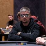 Ein Mann sitzt an einem Pokertisch und hält zwei Spielkarten in der Hand, während er konzentriert auf die Gemeinschaftskarten schaut. Um ihn herum stehen andere Spieler und beobachten gespannt