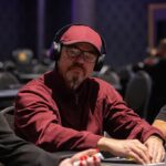 Ein grüner Pokertisch mit Spielkarten und Pokerchips, umgeben von Personen, die ernsthaft spielen.